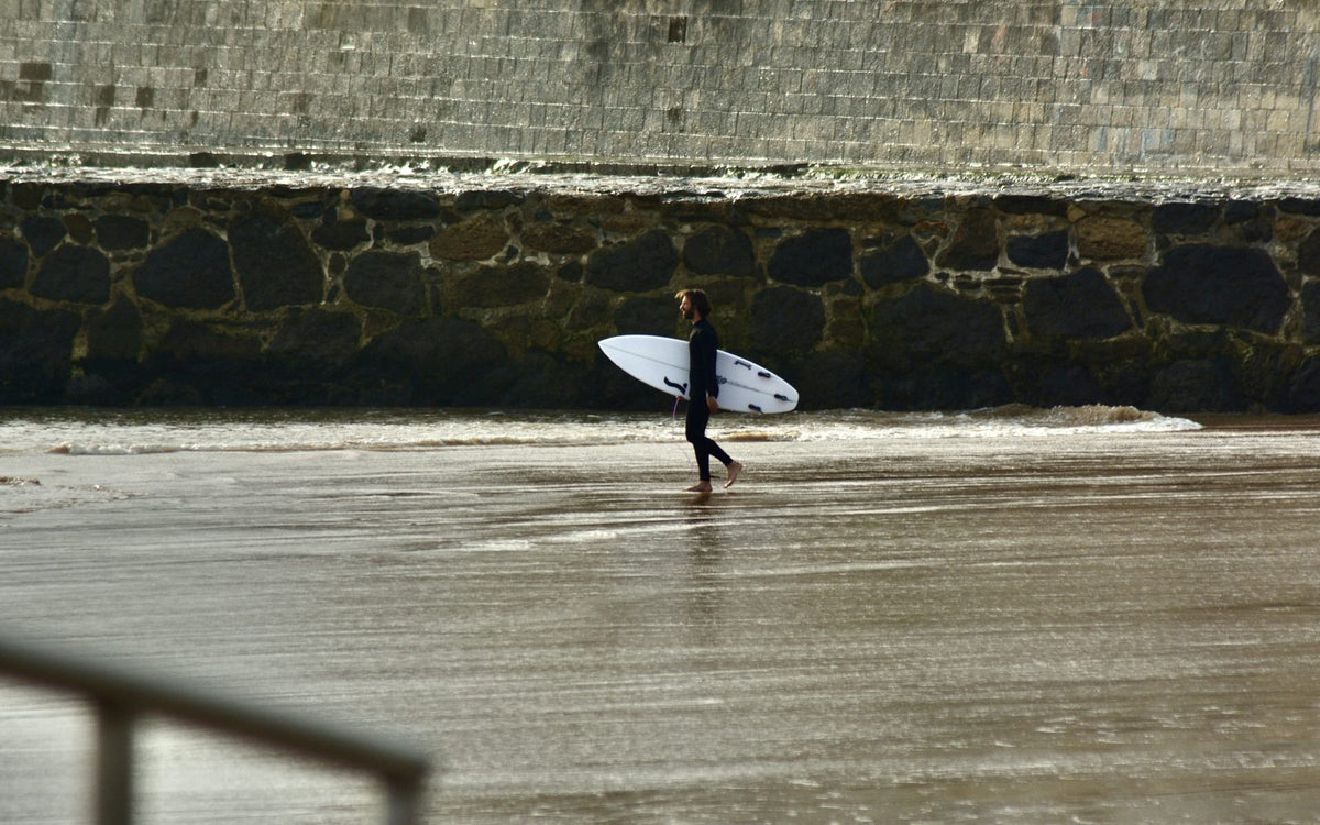 Surfer in a wetsuit carrying a white shortboard across wet sand at the shoreline, sea wall behind