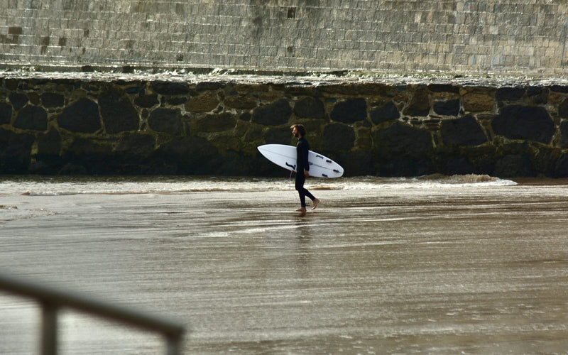Surfer in a wetsuit carrying a white shortboard across wet sand at the shoreline, sea wall behind
