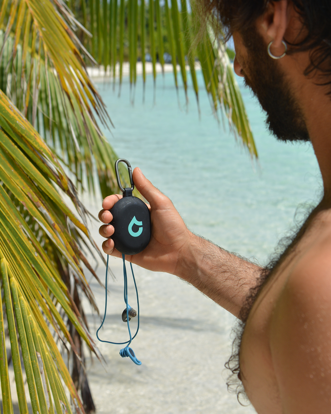 Man holding SEAR Plugs waterproof earplugs case with leash on tropical beach, designed for swimming, surfing and protection against swimmer’s ear