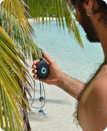 Person holding a black SEAR earplug case with clip near a tropical beach.